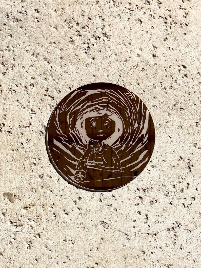 May include: A circular, brown metal coaster featuring a stylized illustration of a girl with large eyes, surrounded by swirling lines. The coaster is placed on a textured, light-colored surface.