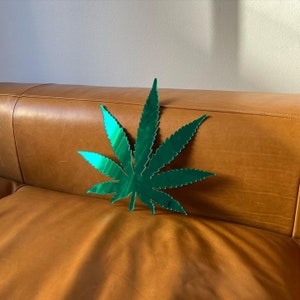 May include: A green mirrored leaf shaped like a marijuana plant on a brown leather sofa.