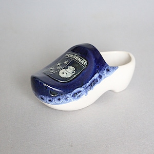 May include: A white ceramic clog with a blue glaze and a coat of arms featuring a pitcher and stars. The coat of arms is labeled "KURSENAI".