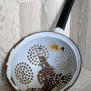 Peut inclure: Une passoire en émail blanc avec une poignée noire et un intérieur rouillé. La passoire est ronde et a un fond perforé.