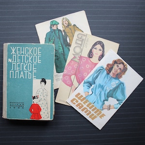 Op de afbeelding: Een verzameling vintage naaipatronenboeken en tijdschriften. Het boek links is lichtblauw met Russische tekst en illustraties van een vrouw en een kind. Verschillende tijdschriften bevatten mode-illustraties.