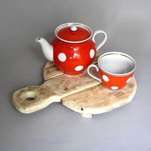 May include: A red teapot and teacup set with white polka dots. The teapot has a white spout, handle, and lid. The teacup has a gold rim. The set is on a wooden serving board.