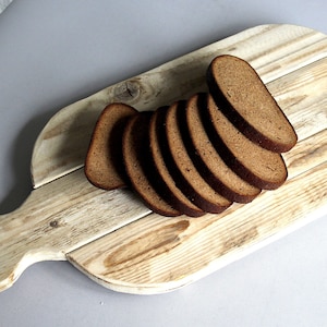 May include: A wooden chopping board with a handle, holding several slices of dark brown bread. The board has a natural wood grain pattern and a light, weathered finish. The bread slices are neatly arranged, suggesting a rustic aesthetic.