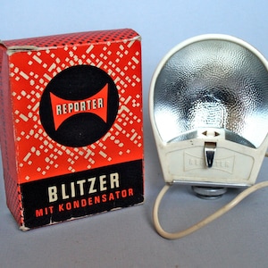 Peut inclure: Un flash blanc avec un réflecteur argenté et le mot "REPORTER" imprimé sur le côté. Le flash est relié à un cordon blanc et est posé sur une surface blanche. Le flash est dans sa boîte d'origine, qui est rouge avec des pois blancs et les mots "BLITZER MIT KONDENSATOR" imprimés sur le devant.