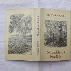 Può includere: Un libro aperto con una copertina beige chiaro con illustrazioni in bianco e nero di alberi e paesaggi. Il dorso mostra il titolo "Skruzdėlynas Prūsijoje" e il nome dell'autore, "Juozas Aputis."