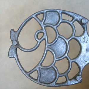 Może przedstawiać: Srebrny metalowy przyrząd kuchenny w kształcie ryby z ozdobnym wzorem. Przyrząd ma otwór w środku i mały okrągły otwór z boku. Przyrząd jest oznaczony "WEDGE" z boku.