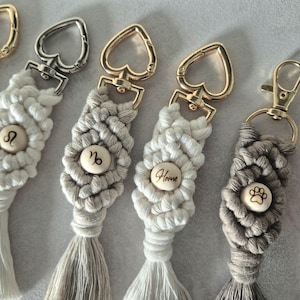 Peut inclure: Porte-clés en macramé avec des attaches dorées et argentées. Les porte-clés présentent des motifs tissés de différentes couleurs neutres, avec des boutons en bois. Certains boutons comportent des symboles du zodiaque, une empreinte de patte ou le mot "Home".