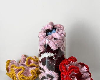 Puede incluir: Una colección de coleteros de ganchillo hechos a mano en varios colores. Un coletero es rosa con ribete amarillo, otro es rojo y rosa, y el resto están en un tarro de cristal transparente. Los coleteros están hechos de hilo suave.