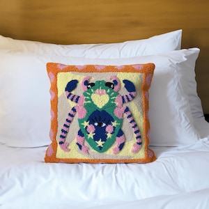 Op de afbeelding: Een vierkant decoratief kussen met een kleurrijk, geborduurd ontwerp. Het kussen heeft een centraal figuur met een blauw gezicht, groen lichaam en roze en blauw gestreepte ledematen, omgeven door een gele achtergrond en een oranje rand. Het ligt op een wit bed.
