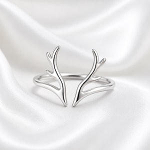 May include: A polished silver-toned open ring featuring two symmetrical, branching antler-shaped ends that curve upwards. The band is smooth and continuous, meeting the base of each antler design, creating a clean, metallic, and reflective appearance.
