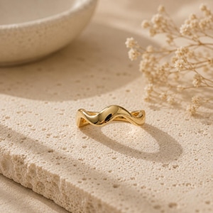 May include: A gold ring with a flowing, wave-like design. The ring is highly polished and reflects light, set against a textured, pale surface. The ring's design is smooth and organic, with a continuous, curved form. A piece of jewellery.