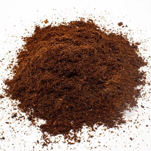 May include: A mound of dark brown, finely textured soil, possibly peat moss or compost, against a white backdrop. The soil is densely packed, with a slightly uneven surface and scattered particles.