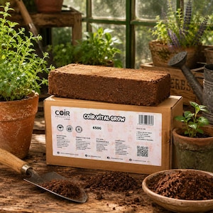 May include: A rectangular brown compressed coir brick sits atop a cardboard box labelled Coir Vital Grow 650g. The box features printed instructions, icons, and a QR code. The product is positioned on a wooden surface with a metal trowel and soil.