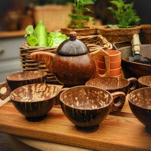 May include: A set of six brown coconut shell teacups and a brown coconut shell teapot with a wooden handle. The cups and teapot are arranged on a wooden surface.