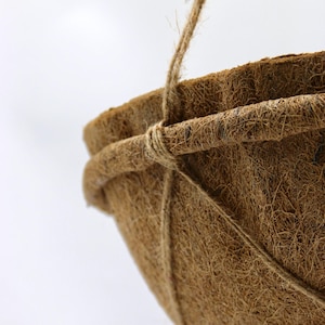 Op de afbeelding: Een bruine, komvormige hangende plantenbakvoering gemaakt van vezelig kokosvezelmateriaal. De plantenbak heeft een dikke, verstevigde cirkelvormige rand en wordt opgehangen aan dunne, natuurkleurige touwtjes die stevig rond de bovenrand zijn geknoopt.