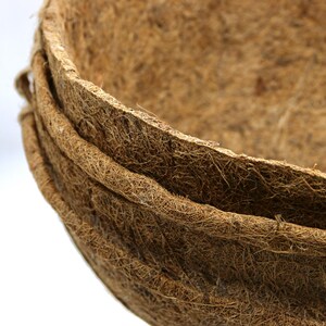 Op de afbeelding: Een stapel van drie ronde, komvormige plantenbakken gemaakt van natuurlijke bruine kokosvezels. De plantenbakken hebben een grove, vezelige textuur met een zichtbare geweven structuur, die een diepe, open komvorm vormen voor aarde en planten.