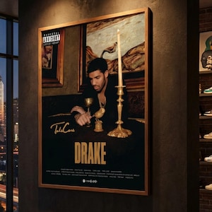 May include: A framed poster featuring Drake, with the artist's name prominently displayed in gold lettering. The image includes a portrait of Drake, a candle, and a golden goblet. The poster is displayed in a modern living space with a city skyline view.