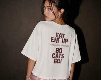 May include: A white short-sleeved t-shirt featuring dark red textured lettering on the back that reads EAT EM' UP, Texas State University, and GO CATS GO! The shirt is paired with light pink satin shorts with a visible hemline and elastic waist.