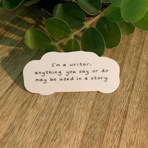 May include: A white sticker in the shape of a cloud with the text "I'm a writer, anything you say or do may be used in a story." The sticker is on a wooden surface with green leaves in the background.