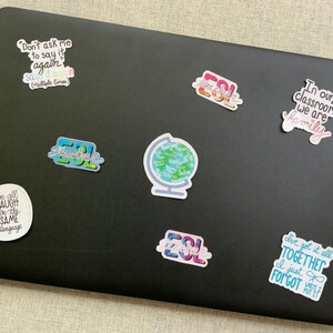 May include: A black laptop with several colourful stickers on it. The stickers feature phrases like "Don't ask me to say it again", "We all laugh in the same language", "ESL Teacher", "In our classroom we are family", and "I just forgot".