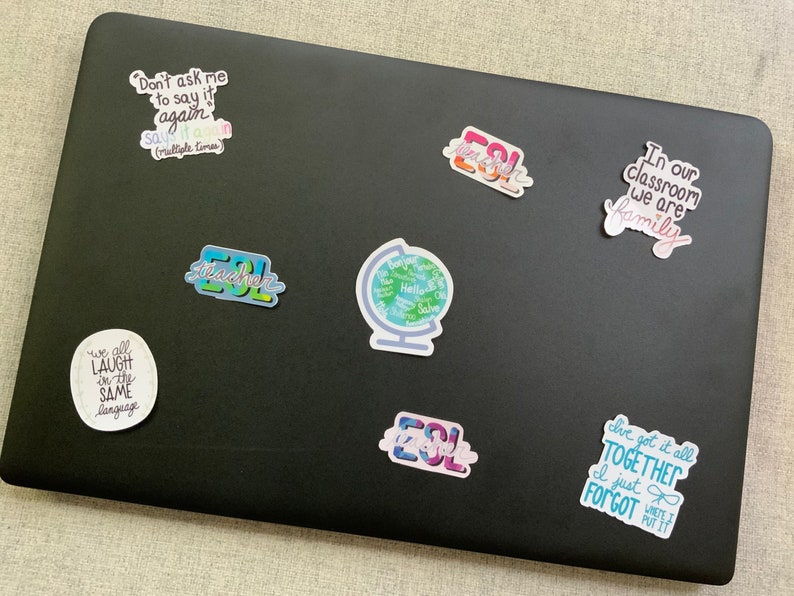 May include: A black laptop with several colourful stickers on it. The stickers feature phrases like "Don't ask me to say it again", "We all laugh in the same language", "ESL Teacher", "In our classroom we are family", and "I just forgot".