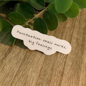 May include: A white sticker in a cloud shape with the text "Punctuation: small marks, big feelings." The sticker is on a wooden surface, with green leaves in the background.