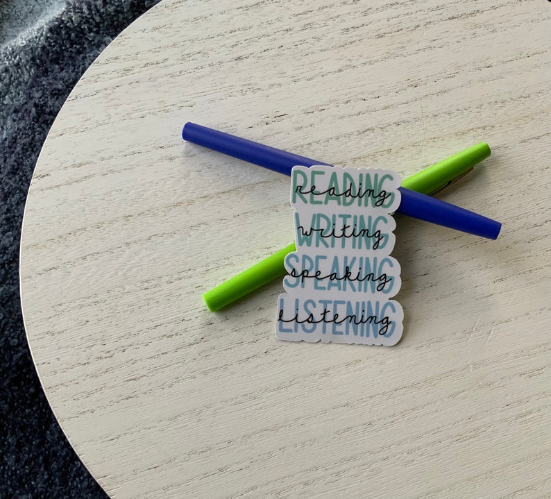 May include: Four stickers with the words "Reading", "Writing", "Speaking", and "Listening" in a blue and green color scheme. The stickers are stacked on top of each other and are on a white wooden surface.
