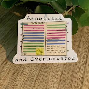 以下が含まれることがあります： 「Annotated and Overinvested」の文字が黒で書かれた白いステッカー。ステッカーには、カラフルな水平線と手書きのメモが2列に並んだデザインがあり、プランナーやノートのページに似ています。