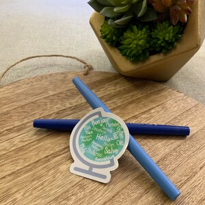 May include: A sticker of a globe with the word "Hello" in the centre and surrounded by greetings in different languages, including "Bonjour", "Merhaba", "Guten Tag", "Shalom", "Salve", and "Konnichiwa".