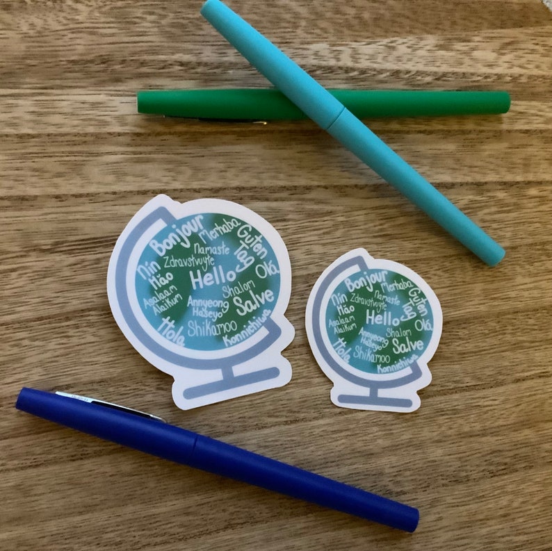 May include: Two round stickers with a globe design. The stickers feature a blue and green globe with the word "Hello" in the centre and surrounded by words for "hello" in different languages. The stickers are on a wooden surface.