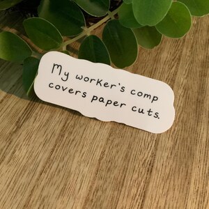 May include: A white sticker with a scalloped edge, featuring the text "My worker's comp covers paper cuts." The sticker is on a wooden surface, with green leaves in the background.