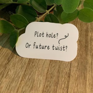 May include: A white sticker in a cloud shape with the text "Plot hole? Or future twist?" in black. The sticker is on a wooden surface, with green leaves in the background.