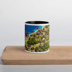 May include: White ceramic mug with a black interior, featuring a detailed map illustration of a medieval village with a river and buildings. The mug is placed on a wooden surface.