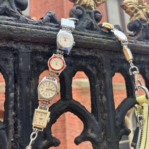 Może przedstawiać: Unikalny dodatek z kolekcją zabytkowych zegarków naręcznych połączonych w pasek. Zegarki mają srebrne i złote koperty z różnymi wzorami tarcz. Widoczna jest również żółta torebka ze srebrnym zamkiem błyskawicznym i czarną metką.