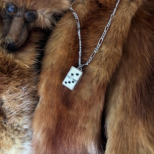 Könnte beinhalten: Eine silberne Kettenkette mit einem weißen Domino-Anhänger. Die Kette liegt auf einer braunen Pelzfläche.