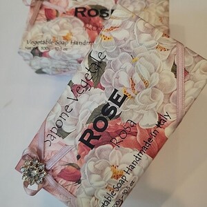 Pode incluir: Duas barras de sabonete vegetal artesanal, cada uma embalada em papel floral com um desenho de rosa. A embalagem inclui o texto "ROSE Rosa" e "Vegetable Soap Handmade in Italy". Cada barra pesa 300g e é amarrada com uma fita rosa e uma joia decorativa.