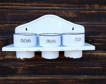 May include: White enamel wall-mounted shelf with three white enamel containers. The containers have black lettering that reads "Пъсъкъ", "Сапунъ", and "Сода".
