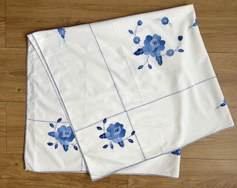 May include: White tablecloth with blue floral embroidery. The tablecloth features embroidered blue flowers and leaves, with blue stitching outlining the edges and design. The cloth is folded, showing the intricate details of the floral pattern.