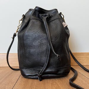 Peut inclure: Sac seau en cuir noir avec fermeture à cordon et longue bandoulière. Le sac a une surface texturée et une quincaillerie dorée. Une petite étiquette avec du texte est attachée sur le côté.