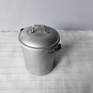Puede incluir: Una olla de aluminio plateada con tapa y asa. La olla tiene una forma ligeramente redondeada y está hecha de metal.