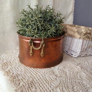 Op de afbeelding: Een koperen plantenbak met messing handvatten, gevuld met een groene kunstplant. De plantenbak staat op een wit gehaakt tafellaken.