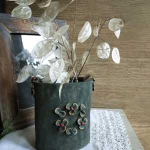 Puede incluir: Un jarrón verde oscuro con una superficie texturizada y adornos florales rojos y dorados. El jarrón está lleno de flores secas, blancas y de papel.