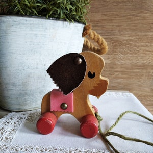 Può includere: Un giocattolo di legno a forma di cane da tirare, con orecchie marroni, una sella rosa e ruote rosse. Il giocattolo ha una corda verde attaccata.