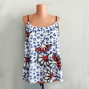 May include: A white camisole with a blue polka dot pattern and red floral designs. The top has thin black straps and a scoop neckline. The floral design has black outlines and red and white petals. The camisole is displayed on a mannequin.