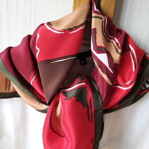 May include: A red, brown, green, and beige silk scarf with an abstract print. The scarf is tied around a white shirt.