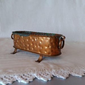 Puede incluir: Un pequeño macetero de cobre martillado con dos asas y cuatro patas. El macetero tiene una pátina verde en el interior.