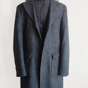 May include: A dark gray herringbone pattern overcoat with a black zip-up inner layer and a high, ribbed collar. The coat has two front pockets and a single-button closure. The coat is displayed on a wooden stand.