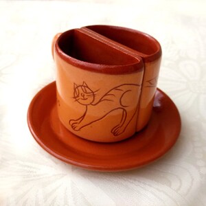 以下が含まれることがあります： 猫のデザインと分割されたトップを持つテラコッタ製のコーヒーカップ。カップは、それに合うテラコッタ製のソーサーの上に置かれています。