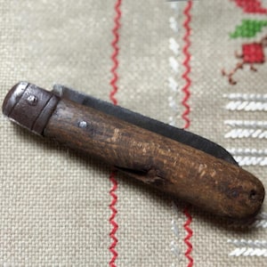 May include: A small, vintage folding knife with a wooden handle and a metal blade. The knife is closed, showing the wooden handle's grain and the metal components. The knife rests on a beige fabric with red stitching.