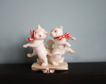 May include: A white porcelain figurine of two cats wearing red and white candy cane bows. The cats are posed in a playful dance, with one cat standing on its hind legs and the other cat sitting on its haunches. The figurine is set on a white base with gold trim.